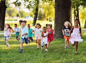 Multiple children running toward camera in grassy park surrounded by trees.