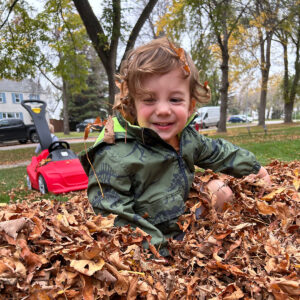 Young child in green jacket sitting in pile of raked leaves in autumn.